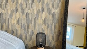 A bedroom with a patterned wallpaper, a round wooden table with a lamp, and a large mirror on the wall. - La Renaissance *** - Collection La 21ème Planche (Bayeux)