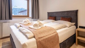A neatly made bed with rolled towels and a blanket, a wooden nightstand, a window with curtains, and a view of mountains.