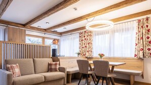 Ein gemütliches Wohnzimmer mit einer Couch, Esstisch und Stühlen. Ein Fenster mit Vorhängen und eine Lampe stehen auf dem Tisch.
