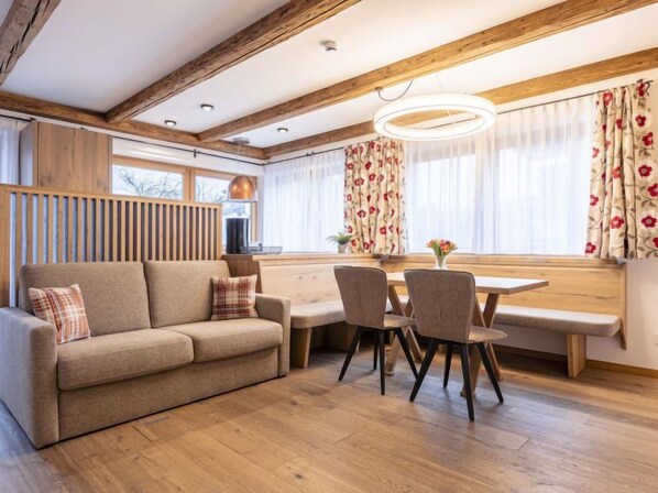 A cozy living room with a sofa, dining table, and chairs. There is a window with curtains and a lamp on the table.