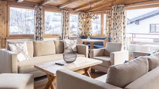 Un intérieur de chalet chaleureux avec un plafond en bois, de grandes fenêtres avec des rideaux, un coin repas et un canapé.
