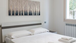 A bedroom with a bed, white bedding, a wall-mounted artwork, and a window with a view of greenery. - Apt24 - Garibaldi 1 in Desenzano del Garda (Desenzano del Garda)