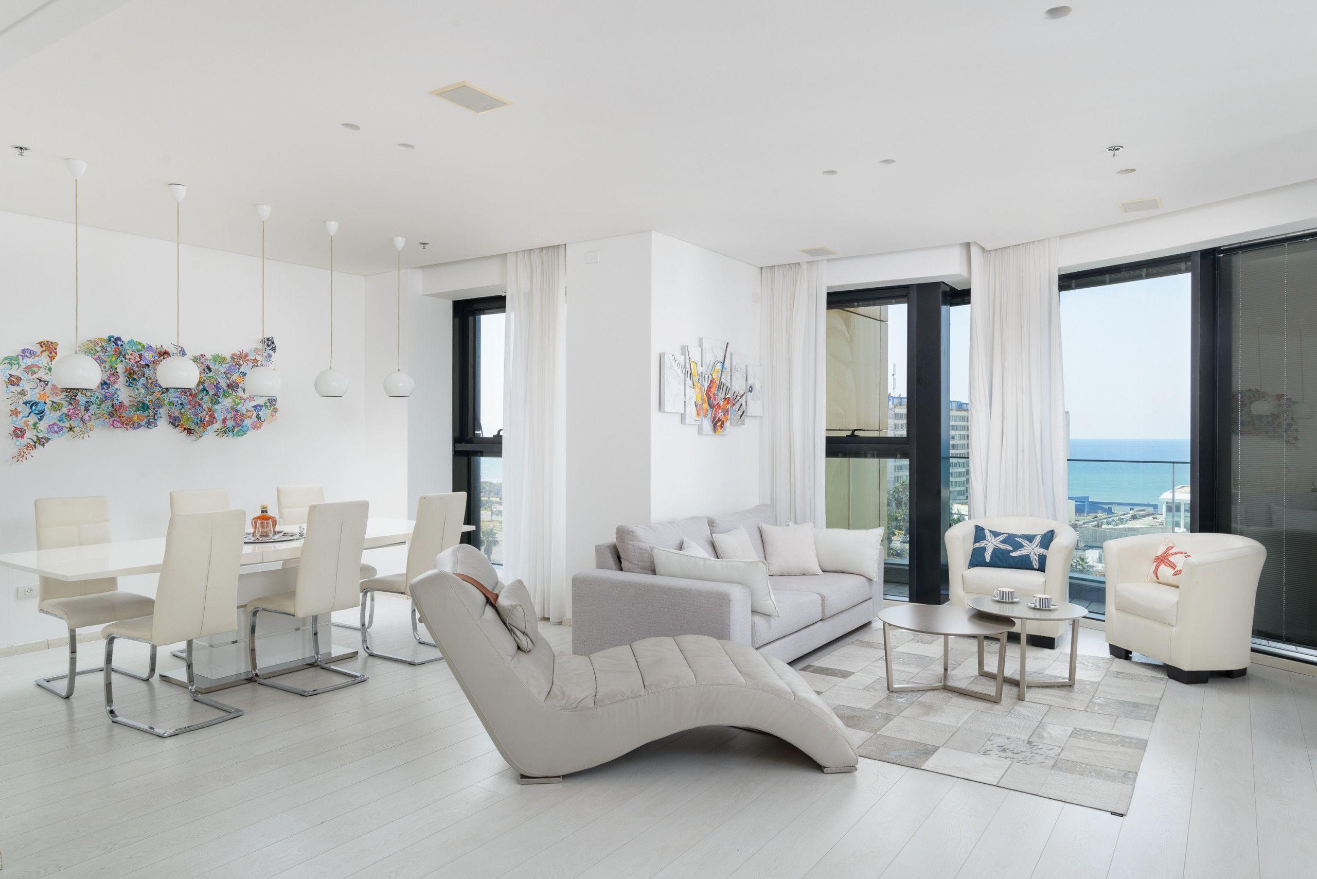A modern living room with a white sofa, a recliner, a dining table with chairs, and a view of the ocean through large windows.