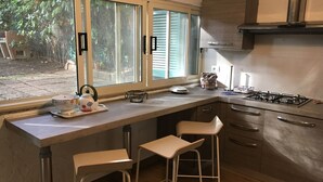 A kitchen with a large window, a gas stove, two bar stools, and a kettle on the counter. - COTTAGE WITH GARDEN AND PRIVATE BEACH (Castiglione della Pescaia)