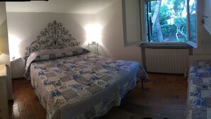 A bedroom with a bed, a bedside table with a lamp, a window with a view of trees, and a radiator. - COTTAGE WITH GARDEN AND PRIVATE BEACH (Castiglione della Pescaia)