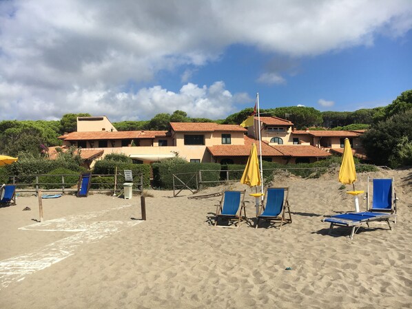A beach with blue lounge chairs and yellow umbrellas, a sandy path leading to a building with red-tiled roofs, and a sign indicating a swimming area.