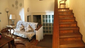 A cozy living room with a sofa, a small table, and a chair. There is a staircase leading to an upper floor. - COTTAGE WITH GARDEN AND PRIVATE BEACH (Castiglione della Pescaia)