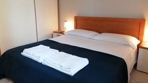 A neatly made bed with white linens and a dark blue bedspread, a bedside lamp, and a small bedside table with a remote control.