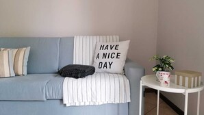 A light blue sofa with striped pillows, a cushion with "HAVE A NICE DAY" text, a small white side table with a plant and a decorative item, and a tiled floor.