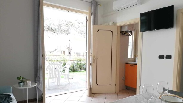 A room with a dining table set for four, a TV mounted on the wall, and a kitchen area visible through an open door.
