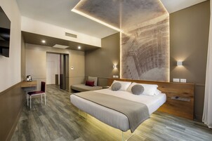 A modern hotel room with a large bed, a desk, and a chair. There is a wall-mounted TV and a large window.