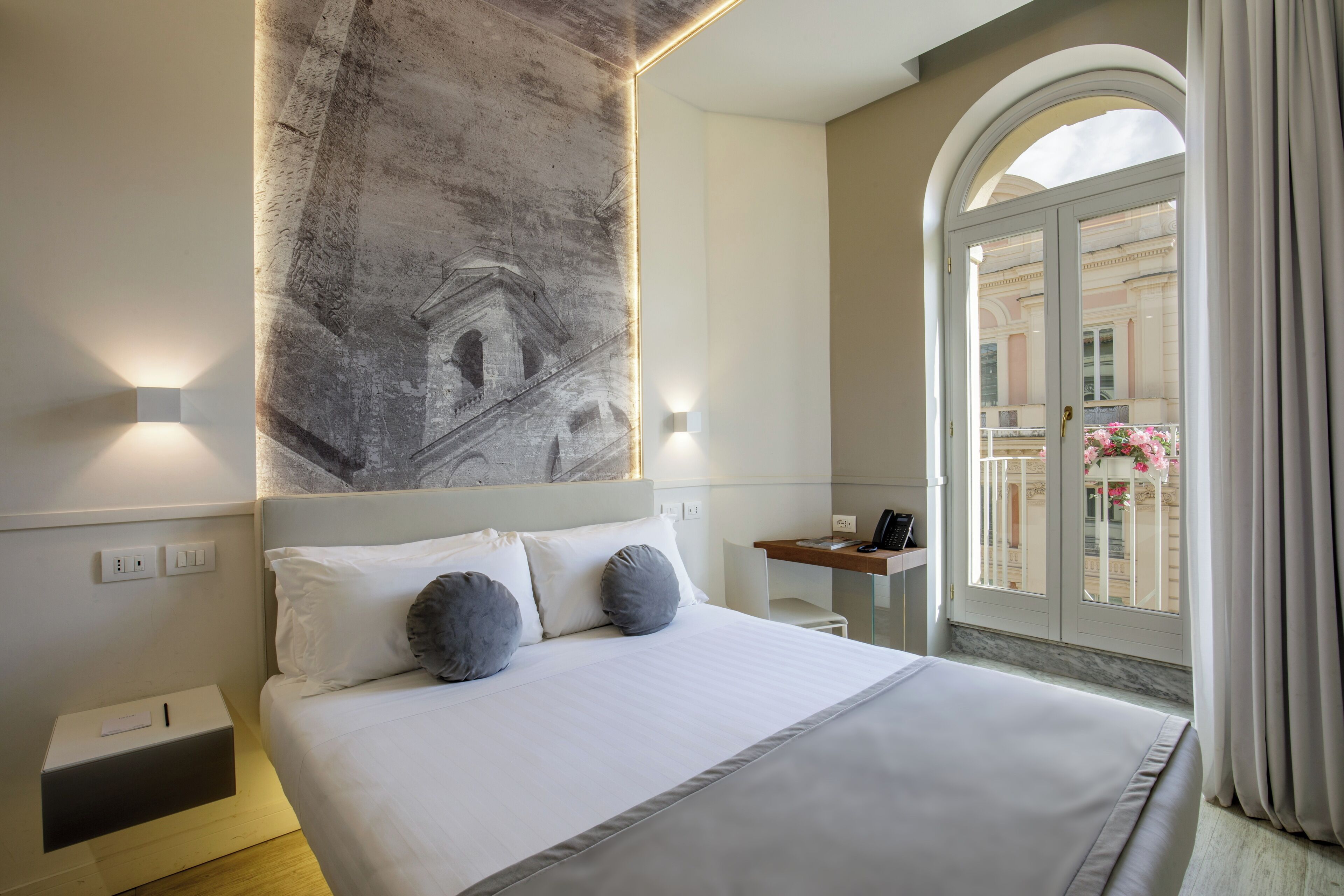 A modern hotel room with a large bed, a desk, and a view of a balcony with flowers.