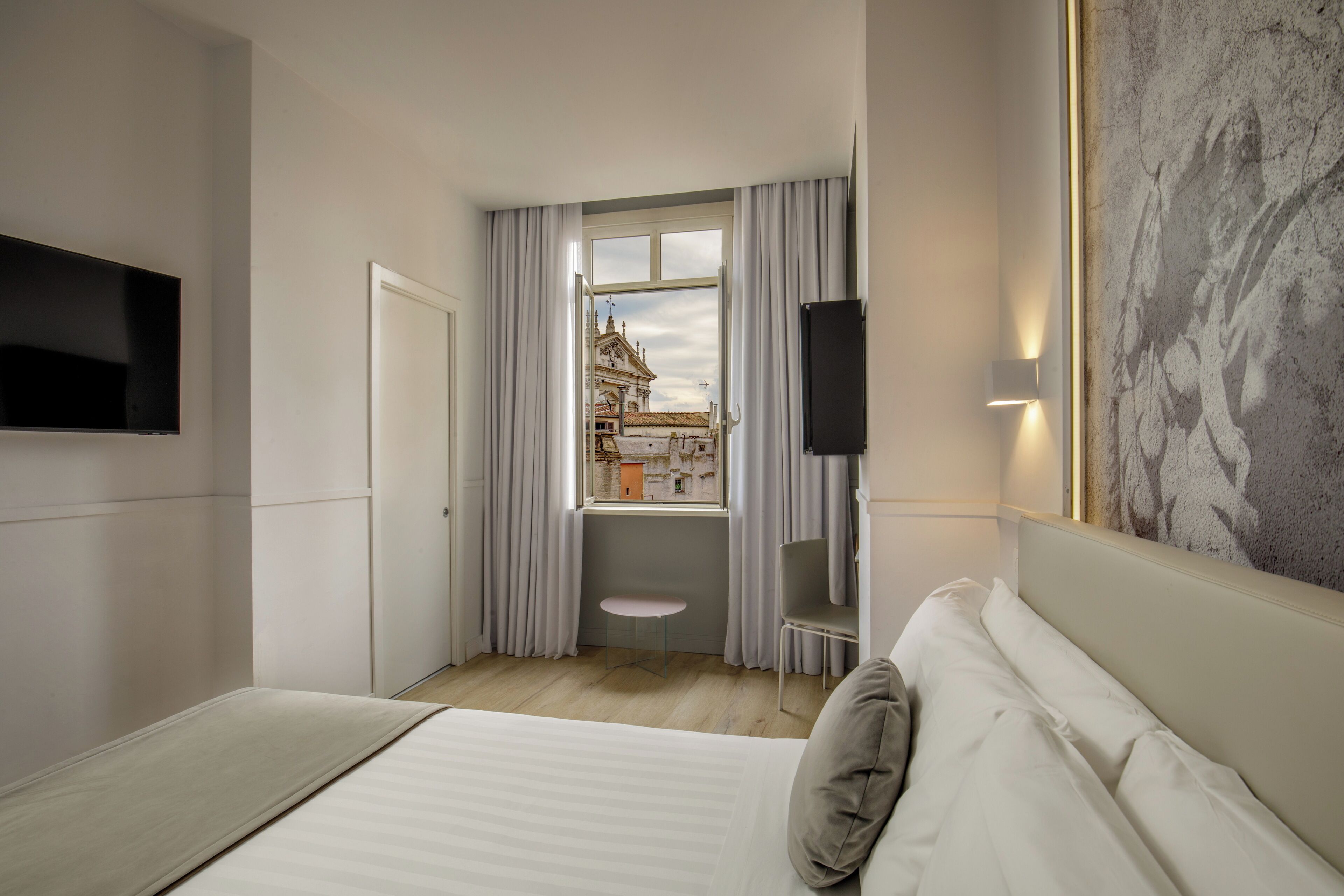 A modern hotel room with a bed, a TV, a small table, and a view of a building through a window.