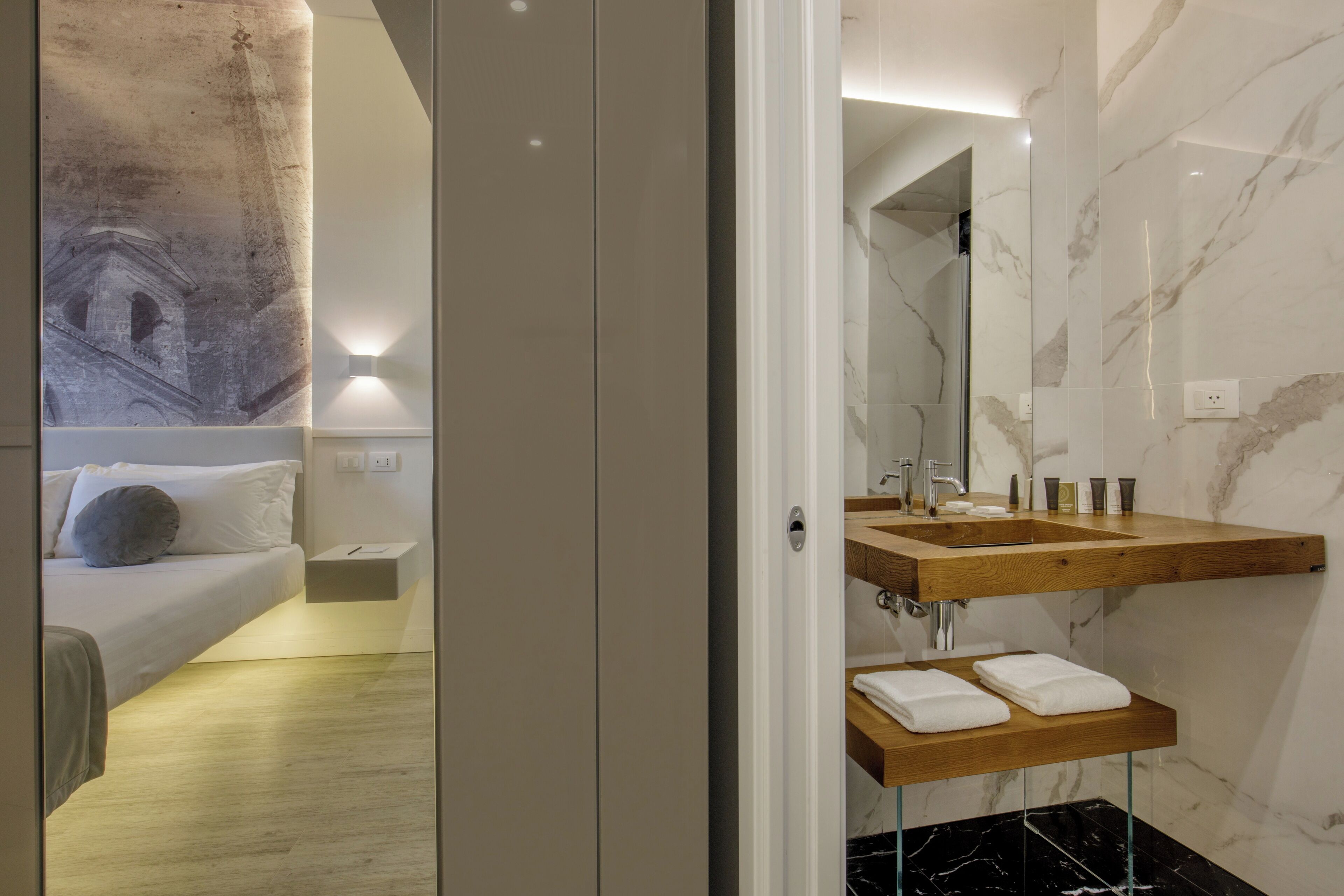 A modern hotel room with a bed, a wooden vanity with a mirror, and a marble shower area.