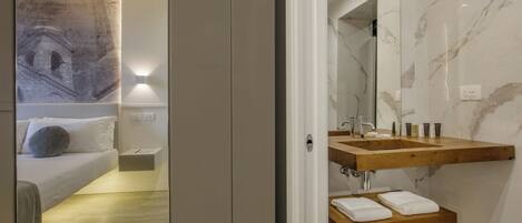 A modern hotel room with a bed, a wooden vanity with a mirror, and a marble shower area.