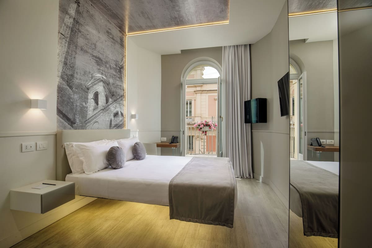 Moderni hotellihuone, jossa on suuri sänky, taulutelevisio, yöpöytä ja näkymä kaupunkiin holvikaaren muotoisesta ikkunasta.