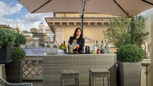 Un área de bar con mobiliario de mimbre, un cantinero preparando una bebida y una vista de edificios históricos al fondo.