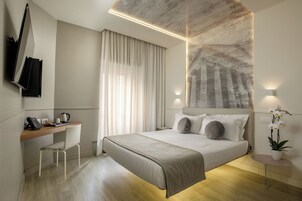 A modern hotel room with a bed, a desk, a chair, a small table, and a potted plant.
