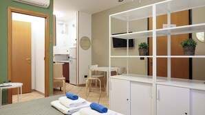 Une chambre d’hôtel avec un lit, un petit bureau, une chaise, une télévision et une étagère avec des plantes.