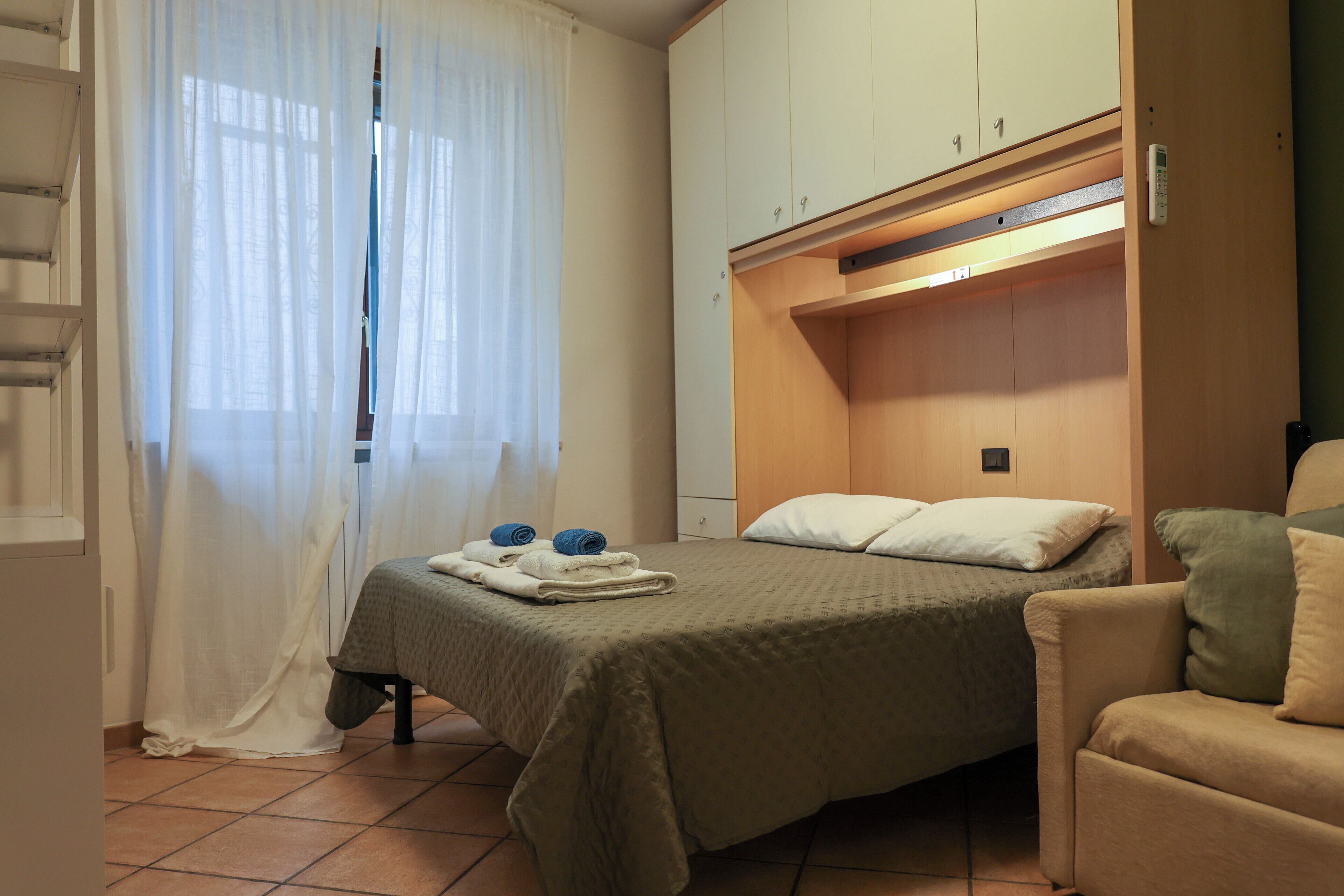 Ein Schlafzimmer mit Bett, Sessel, Kleiderschrank und einem Fenster mit Vorhängen.