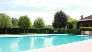 A rectangular swimming pool with a tiled deck, surrounded by greenery and a building with a tiled roof.