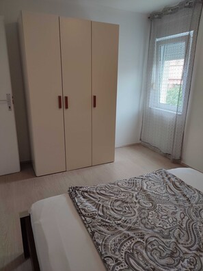 A bedroom with a large wardrobe, a bed with a patterned bedspread, and a window with a view of greenery. - Apartment Horvat (Dobrinj)