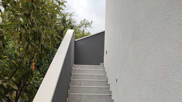 Un escalier en béton, doté d’une rampe blanche, mène à un bâtiment dont la façade présente un aspect texturé.