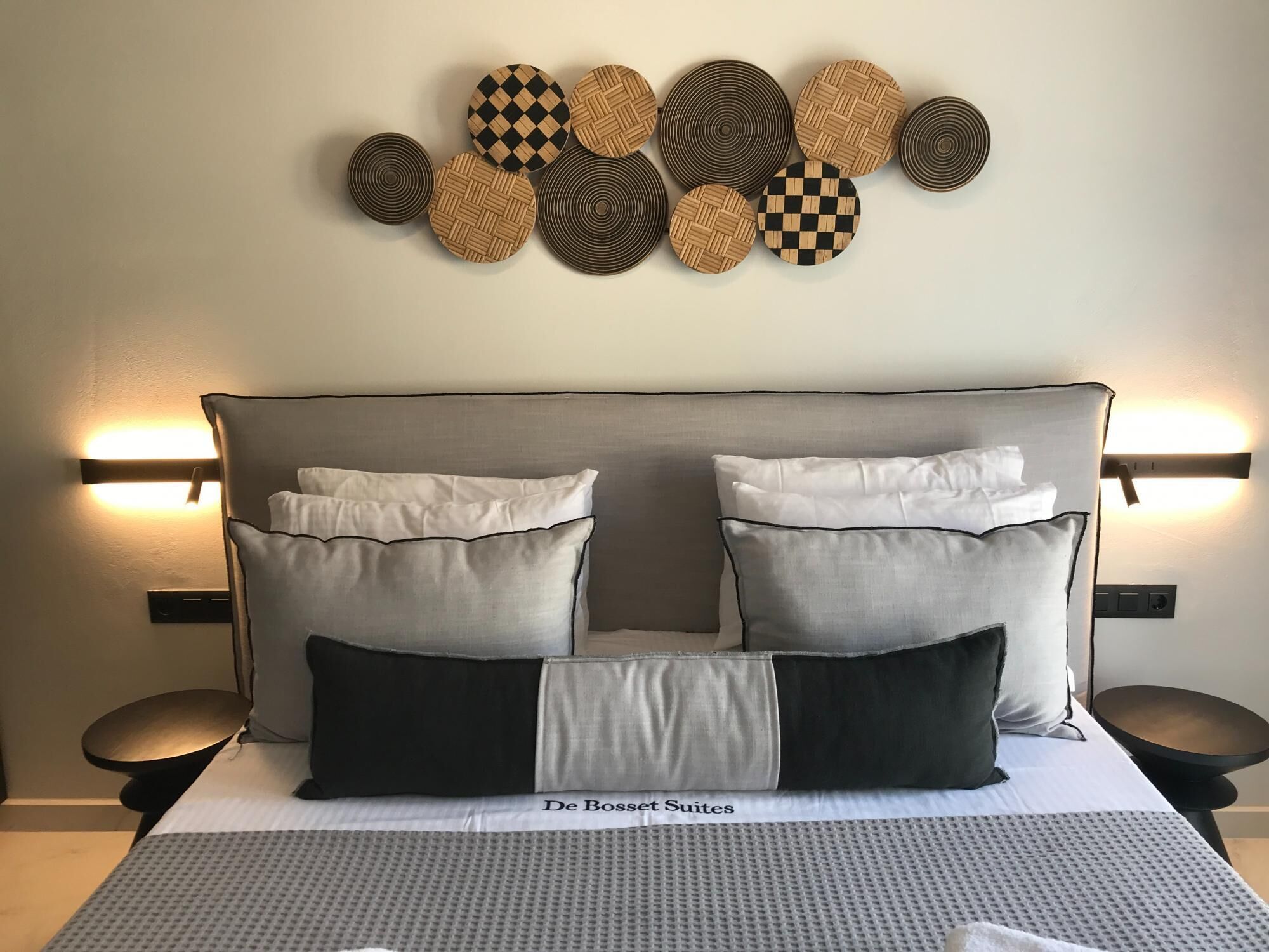 A bed with a grey headboard, white and black pillows, and a grey patterned bedspread. Above the bed, there is a wall-mounted light fixture and a decorative wall art piece.
