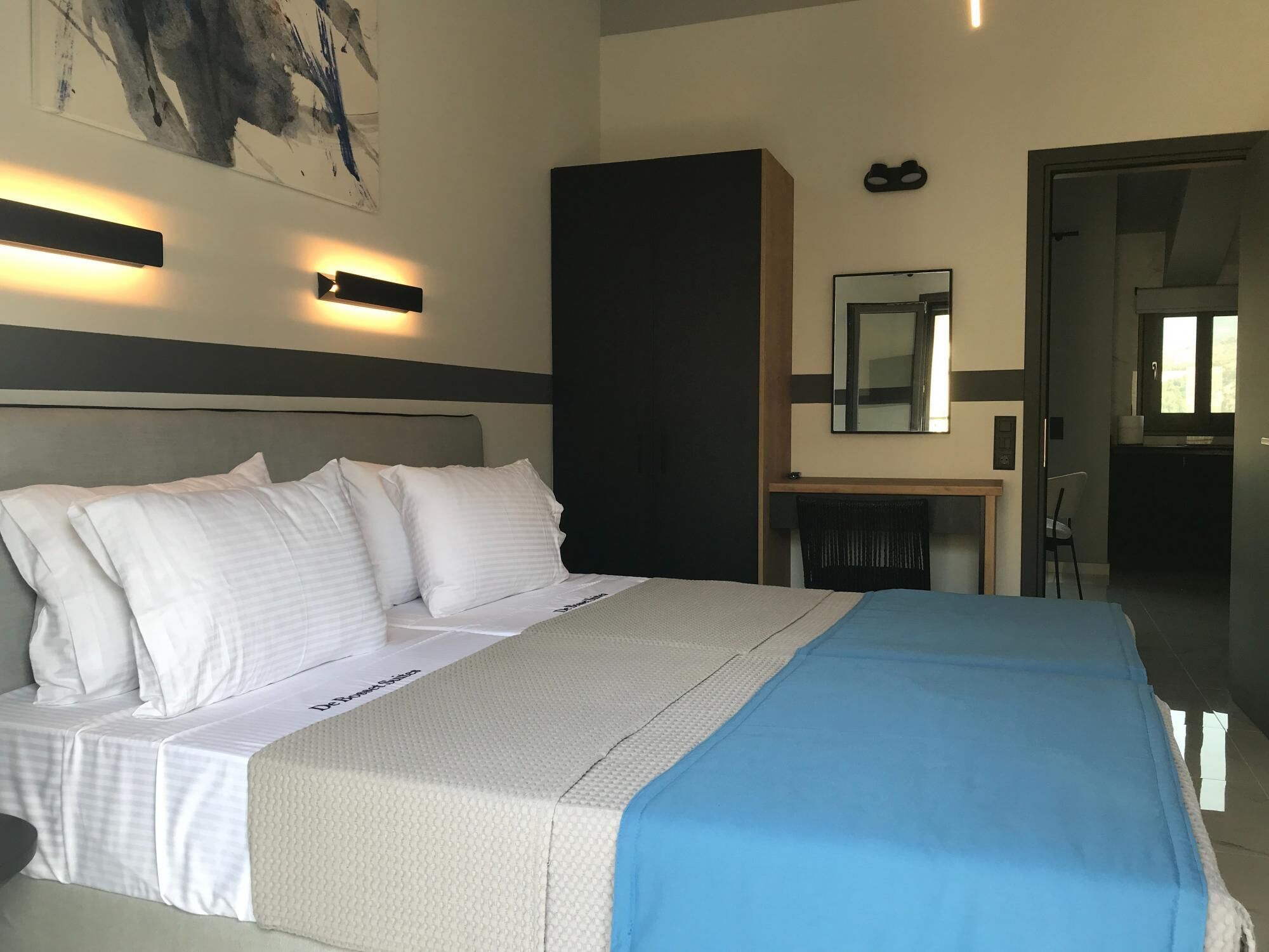 A modern hotel room with a large bed, a desk, a chair, a mirror, and a painting on the wall.