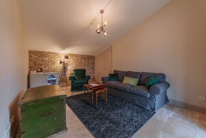 Una sala de estar con un sofá gris, un sillón verde, una mesa de centro de madera, una alfombra negra y una pared de ladrillo.