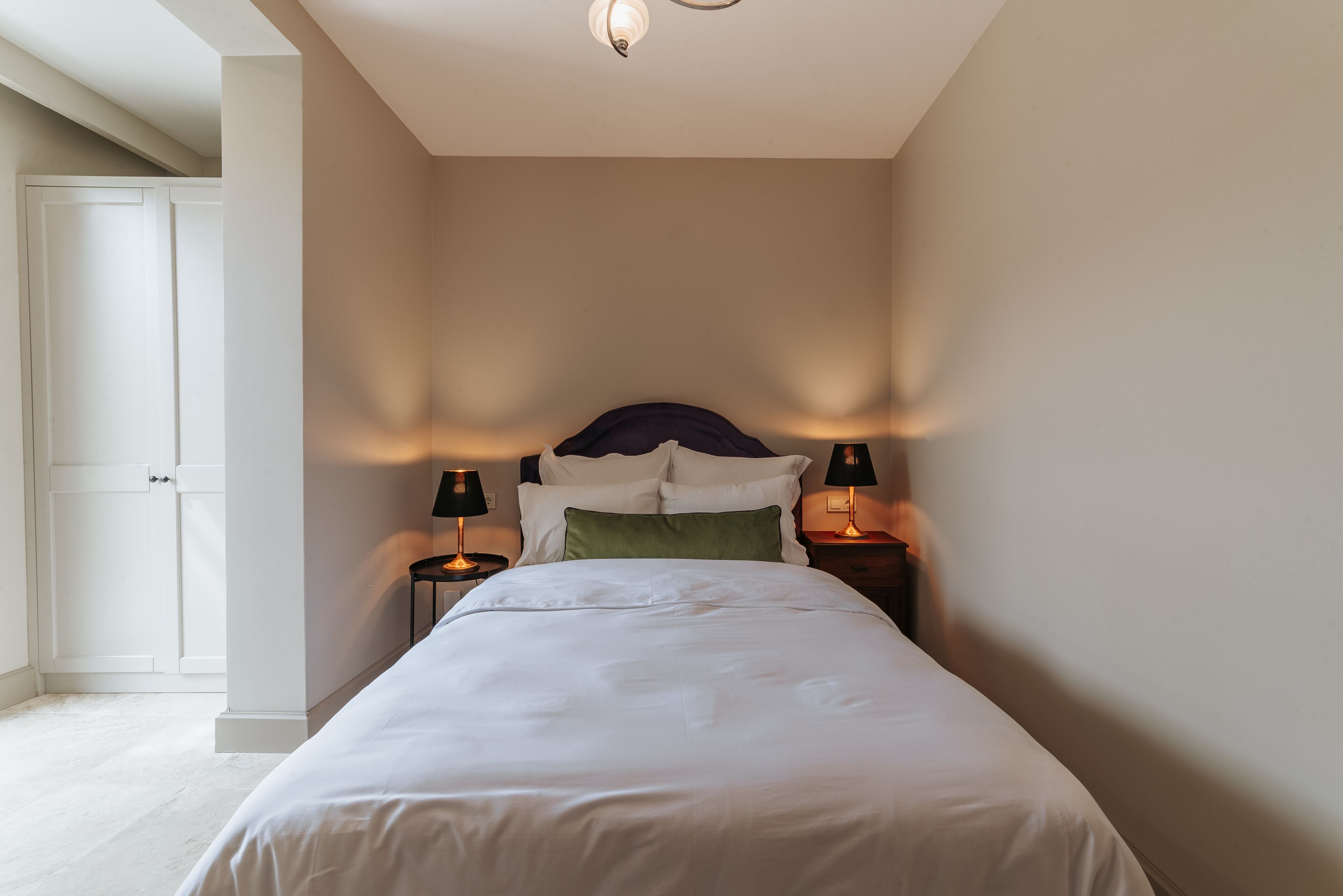 A bedroom with a bed, two bedside lamps, a nightstand, and a door leading to another room.
