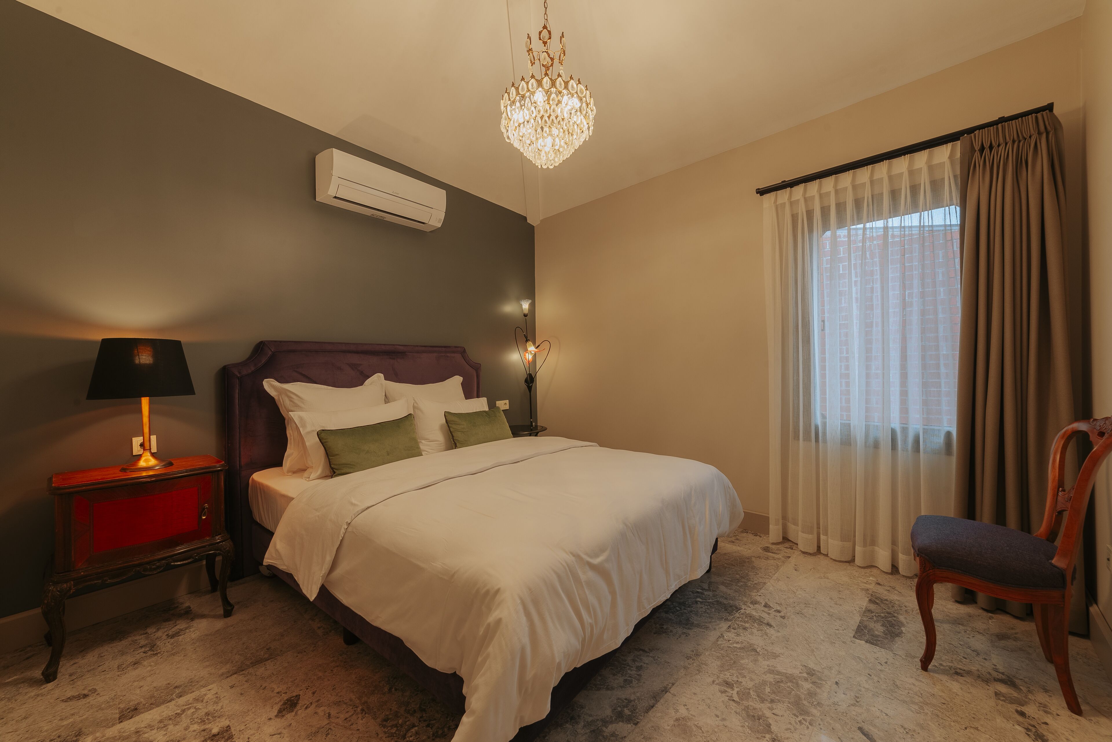 A bedroom with a bed, a nightstand, a lamp, a chair, a chandelier, and a window with curtains.