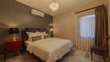 A bedroom with a bed, a nightstand, a lamp, a chair, a chandelier, and a window with curtains.
