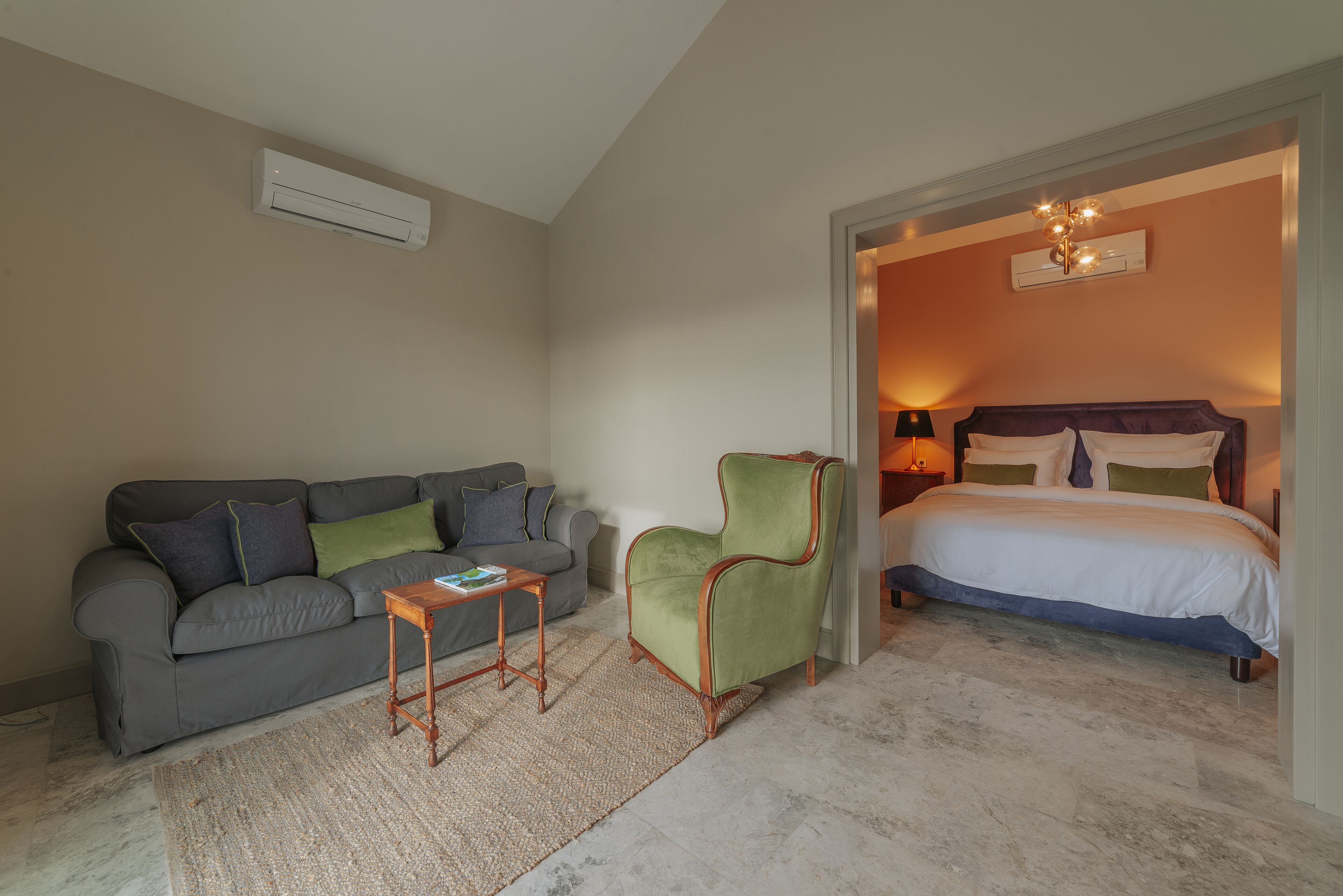 A room with a grey sofa, a green armchair, a small wooden table, and a bed with a white duvet.