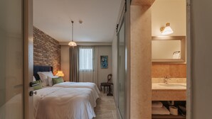 A hotel room with a brick accent wall, a bed with white bedding, a bedside table with a lamp, a window with curtains, a painting on the wall, and a bathroom with a sink and mirror. - oDam Otel (Urla)