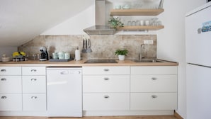Een moderne keuken met witte kastjes, een vaatwasser, een spoelbak en een fornuis. Boven het aanrecht bevinden zich houten planken met bekers en een plant.