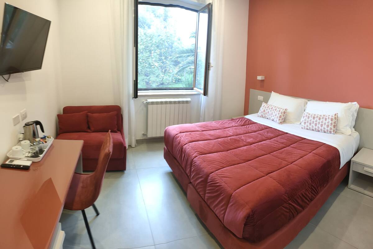 A hotel room with a large bed, a desk with a chair, a TV mounted on the wall, and a window with a view of trees.