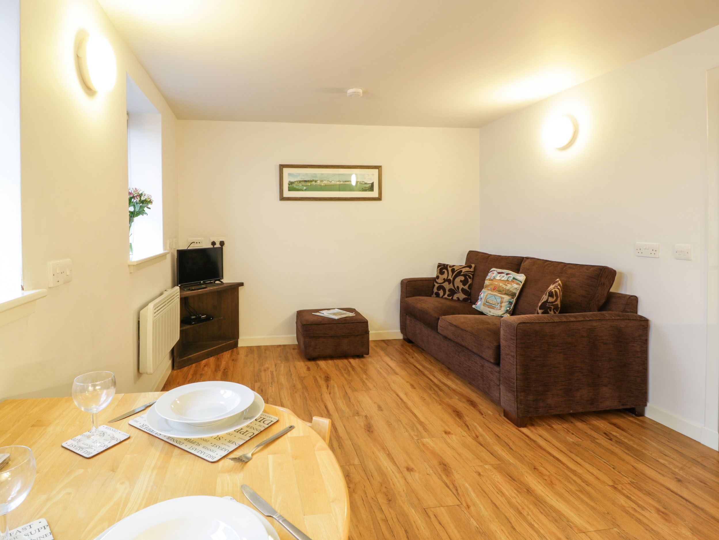 A living room with a brown sofa, a small table with a glass, plates, and cutlery, a television on a stand, and a framed picture on the wall.
