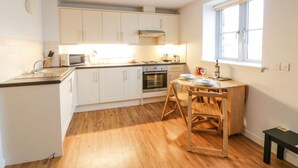 A compact kitchen with white cabinets, a microwave, an oven, and a dining area with a wooden table and chairs. - Tides Reach (Newton Stewart)