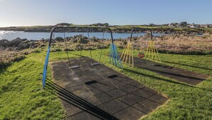A playground with swings and a slide, located near a body of water and grassy area. - Tides Reach (Newton Stewart)