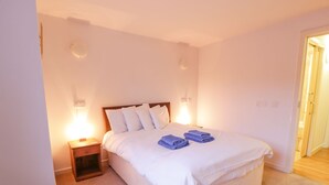 A neatly made bed with white linens and blue towels, a wooden nightstand with a lamp, and a door leading to another room. - Tides Reach (Newton Stewart)