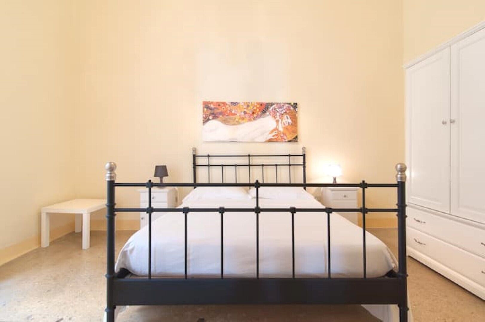 A bedroom with a black metal bed frame, white bedding, a bedside table with a lamp, a nightstand, and a wardrobe.