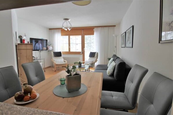 Een moderne woonkamer met een houten eettafel, grijze banken en een televisie aan de muur.