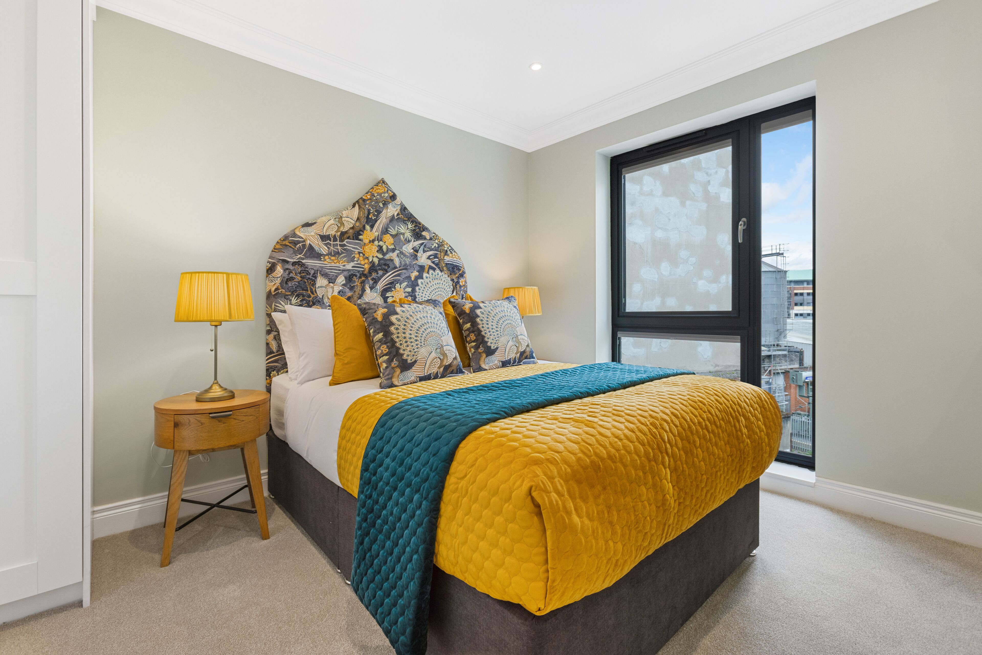 A bedroom with a bed, two bedside tables, a window, and a view of the city.