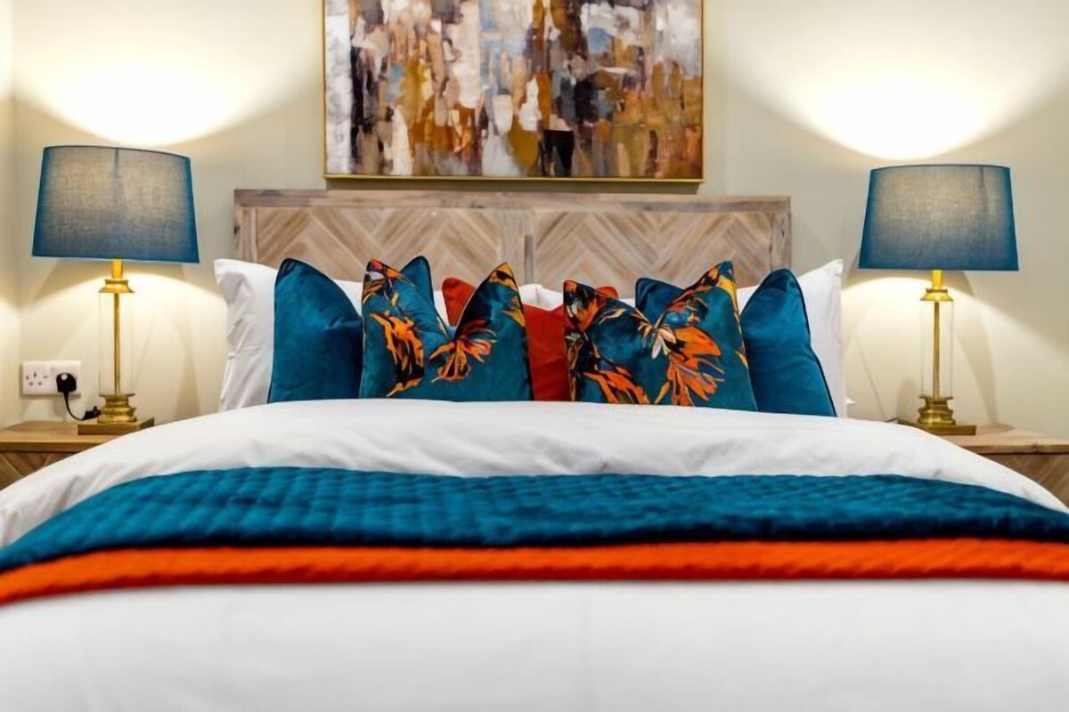 A neatly made bed with a patterned bedspread, two bedside lamps, and a painting above the headboard.