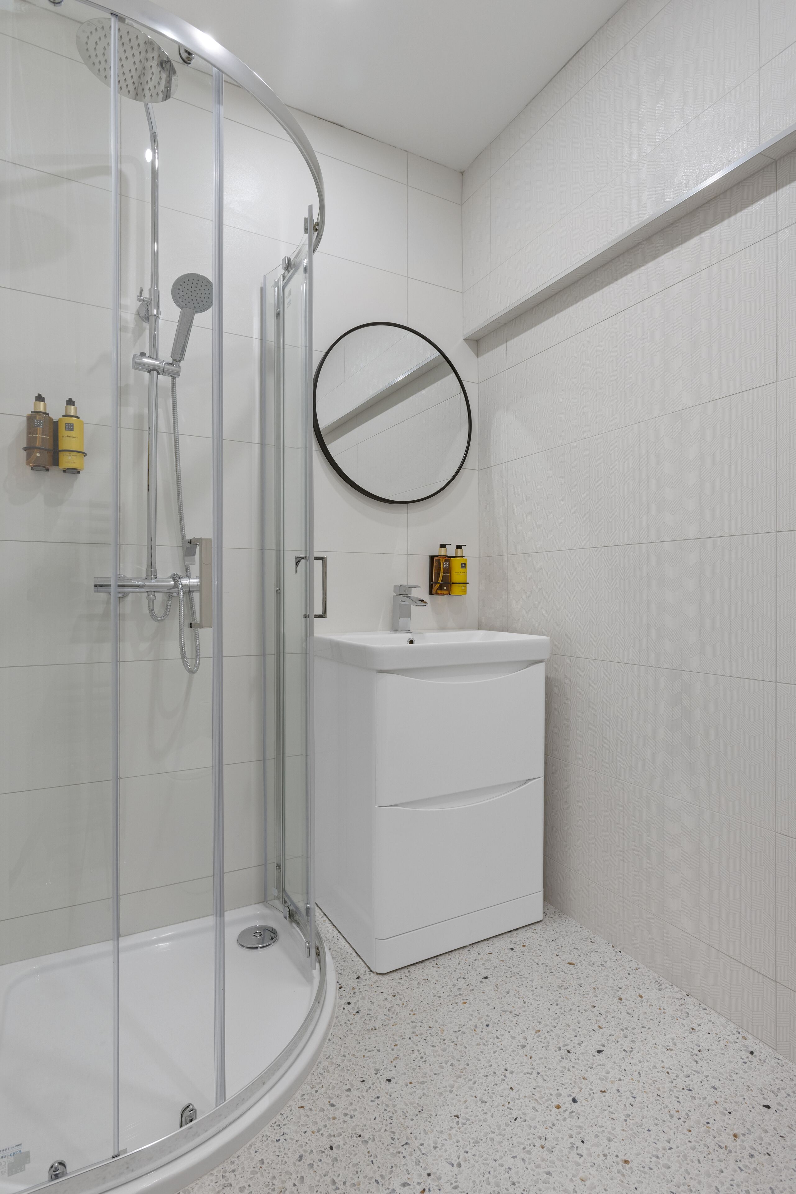 Un baño moderno con cabina de ducha de vidrio, un lavabo blanco con espejo redondo y dos botellas amarillas en la pared.