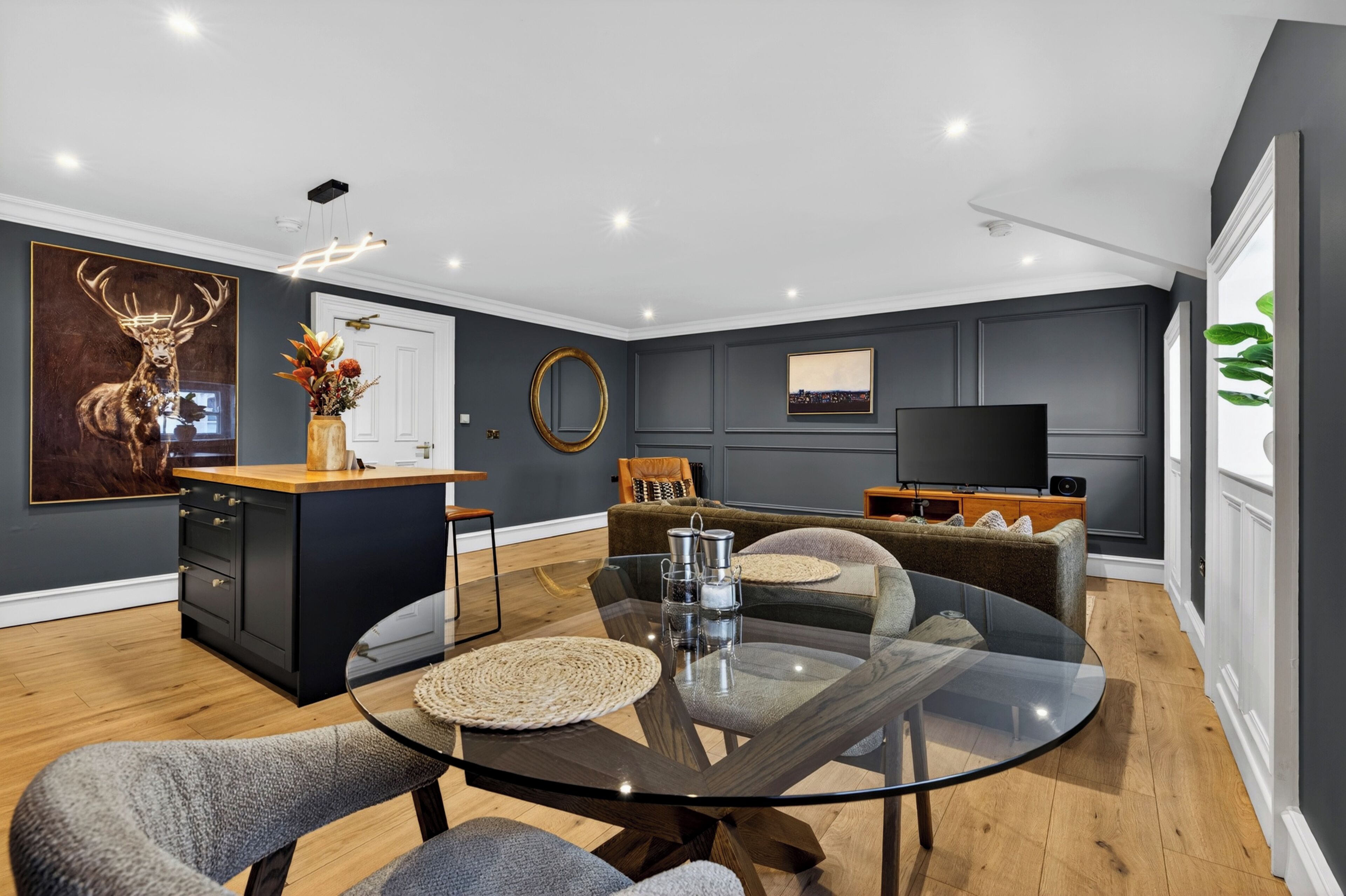 Una sala de estar moderna con una mesa de comedor de vidrio, un sofá, un televisor y una zona de cocina con encimera de madera.