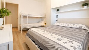 Una habitación con literas, una mesita de noche, una lámpara, una planta en maceta y una estantería con objetos decorativos.