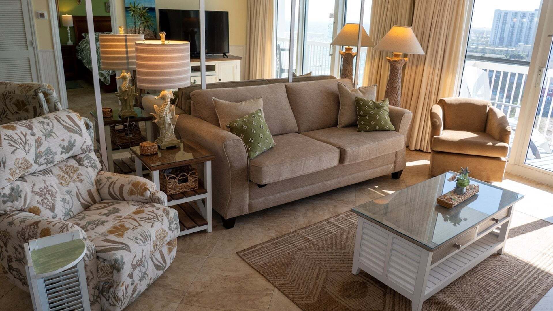 Een woonkamer met een beige bank, een gestreept fauteuil en een glazen salontafel. Er zijn twee vloerlampen, een televisie en een uitzicht op de stad door grote ramen.