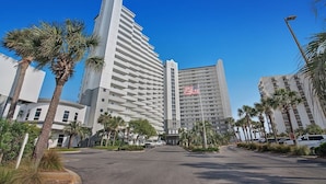 A modern hotel with a prominent American flag, surrounded by palm trees and a clear blue sky. - Pelican Beach 0216 2 Bedroom Condo by Pelican Beach Management (Destin)